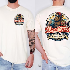 Puede incluir: Camiseta de color crema con un gr&aacute;fico en la parte delantera y trasera. El gr&aacute;fico de la parte delantera muestra el texto "Dad Tax" y un dibujo de Bigfoot. El gr&aacute;fico de la parte trasera es una versi&oacute;n m&aacute;s grande del mismo dise&ntilde;o, con texto adicional.
