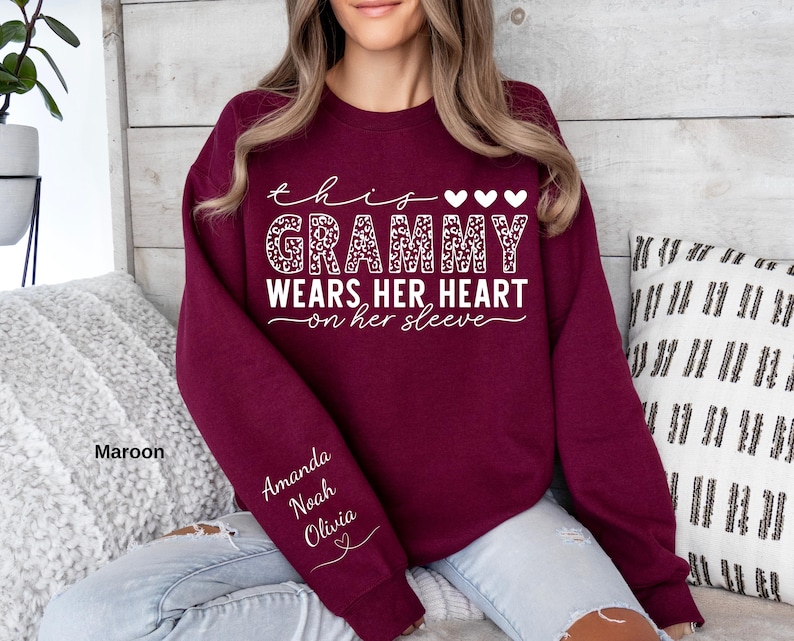May include: Maroon sweatshirt with the text "this Grammy wears her heart on her sleeve" in white script and leopard print. The sleeve has the names "Amanda, Noah, Olivia" in white script. The sweatshirt is a solid maroon color.