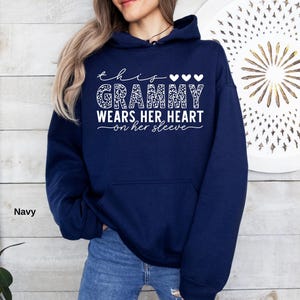 May include: Navy blue hoodie with the text "this grammy wears her heart on her sleeve" in white script. The word "grammy" is in a leopard print pattern. Three white hearts are above the word "grammy".