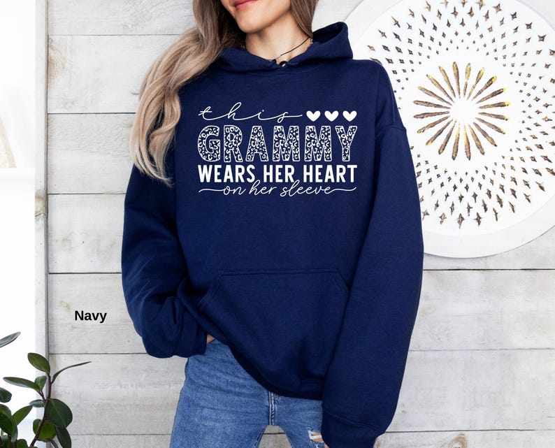 May include: Navy blue hoodie with the text "this grammy wears her heart on her sleeve" in white script. The word "grammy" is in a leopard print pattern. Three white hearts are above the word "grammy".