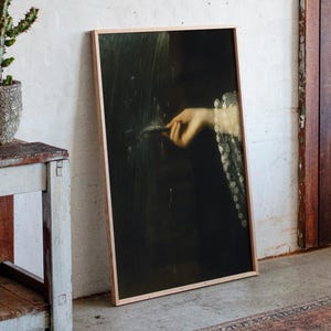 以下が含まれることがあります： 羽を持っている手の古典的な絵画が描かれた額入りのアートワーク。 アートワークは、木のドアの隣の白い壁に立てかけられています。 小さなサボテンが素朴な木製のテーブルの上にあります。