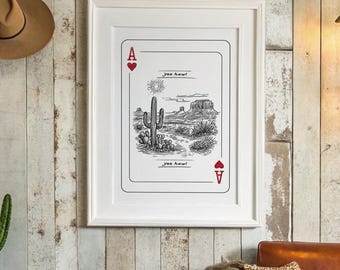 Op de afbeelding: Een ingelijste zwart-wit print van een speelkaart met een woestijnlandschap. De print zit in een witte lijst en hangt aan een rustieke houten muur. Een bruine cowboyhoed en een cactus zijn ook zichtbaar.