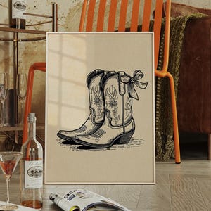May include: A framed print featuring a black and white illustration of cowboy boots. The boots have floral designs and a bow detail. The artwork is displayed on a light-colored background, leaning against a wall in a room with a vintage aesthetic.