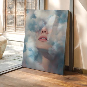 May include: A canvas print featuring a woman's face partially obscured by blue and white clouds. The artwork is displayed indoors, leaning against a wall near a window, with a wooden floor.