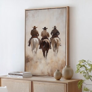 Könnte beinhalten: Ein gerahmtes Gemälde von drei Cowboys, die auf Pferden durch eine staubige Landschaft reiten. Das Gemälde ist in einem hellen Holzrahmen gefasst und hängt an einer weißen Wand über einem Holzschrank.