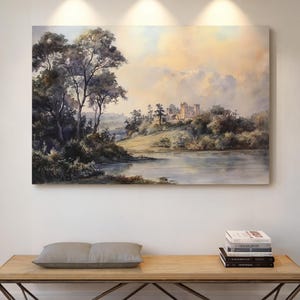 May include: A large landscape painting depicts a serene scene with a lake, trees, and a distant castle under a cloudy sky. Below, a wooden bench with a metal frame holds a pillow and books. Three industrial-style lights illuminate the artwork.