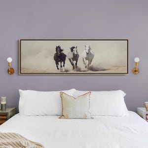 May include: A framed artwork depicting three horses running through a dusty environment. The horses are a dark brown, gray, and white. The artwork is rectangular and hangs above a bed with white linens.