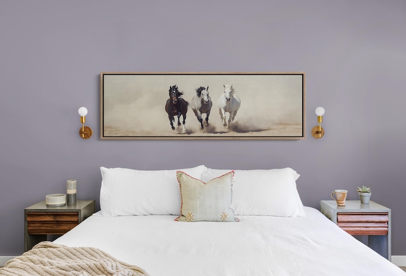May include: A framed artwork depicting three horses running through a dusty environment. The horses are a dark brown, gray, and white. The artwork is rectangular and hangs above a bed with white linens.