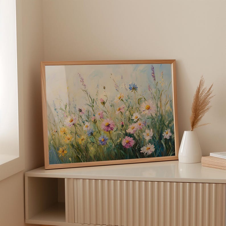 May include: A framed floral painting featuring a field of colorful wildflowers in shades of pink, yellow, blue, and white. The artwork is set in a light brown wooden frame. A white vase with dried grass sits on a cream-colored cabinet.