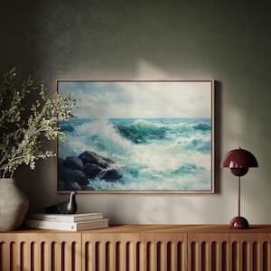 May include: A framed landscape painting depicts a seascape with crashing turquoise waves against dark rocks under a cloudy sky. The artwork is displayed above a wooden cabinet, next to a burgundy table lamp and a vase with greenery.