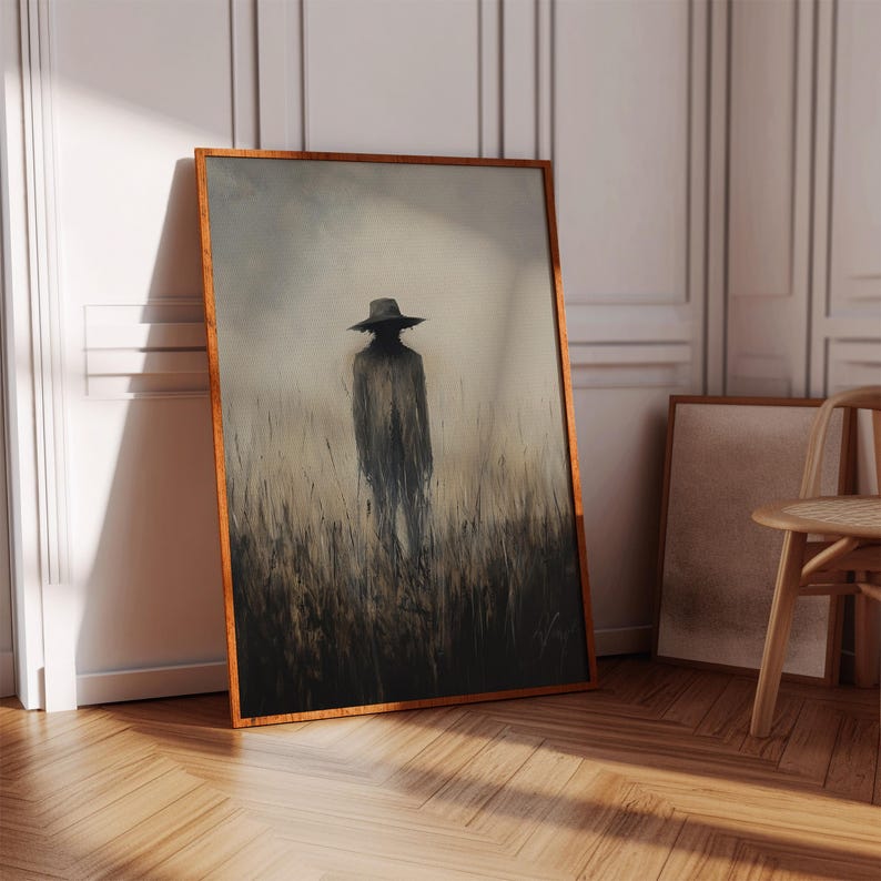 Peut inclure: Une &oelig;uvre d'art encadr&eacute;e repr&eacute;sente une silhouette portant un chapeau dans un champ d'herbes hautes. L'&oelig;uvre a une palette de couleurs sombres et sourdes avec un cadre en bois. L'&oelig;uvre est appuy&eacute;e contre un mur blanc.