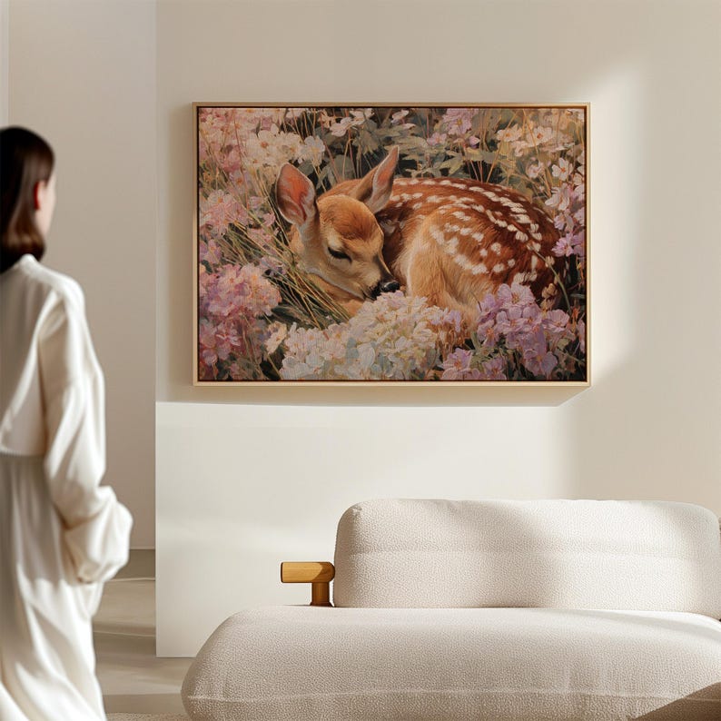 May include: A framed painting depicts a sleeping fawn nestled among pink and white flowers. The fawn has brown fur with white spots. The painting is in a light-colored wooden frame and hangs on a neutral-colored wall.
