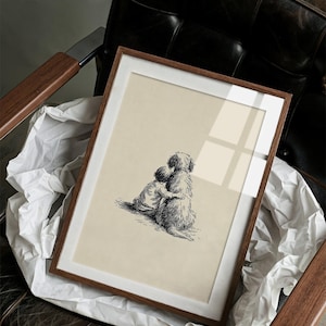 May include: A framed print featuring a black and white illustration of a child hugging a large dog. The artwork has a cream-colored background and is set in a wooden frame with a white mat. The print is resting on white paper inside a leather chair.