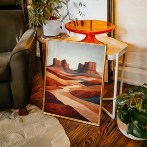 May include: A framed landscape art print featuring a desert scene with layered red, brown, and tan rock formations. The artwork has a gold frame and is displayed on a wooden floor. The color palette includes shades of red, brown, and blue.