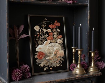 Puede incluir: Una impresión artística enmarcada que presenta un esqueleto de conejo blanco rodeado de flores sobre un fondo negro. La impresión se exhibe en un mueble de madera oscura, con candelabros dorados y velas negras.