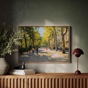 May include: An oil painting in a wooden frame depicts a sunlit street lined with tall trees and buildings. Figures stroll along the path, bathed in warm light. The artwork hangs above a wooden cabinet, complemented by a burgundy lamp and decorative items.