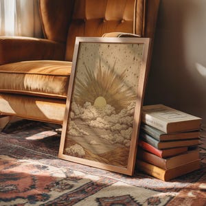 May include: A framed art print featuring a sunburst design with a yellow sun and cloud details. The artwork is in a light brown wooden frame. A stack of books is next to the framed art. The art is on a patterned rug.