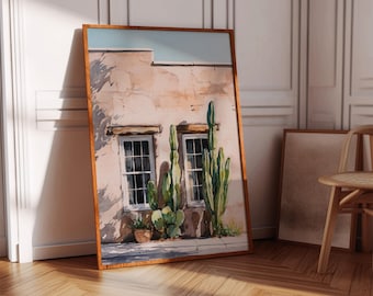 May include: A framed watercolor print of a building with two windows and cacti. The artwork features a light brown building with white-framed windows and green cacti. The frame is a warm brown color, and the artwork is leaning against a white wall.