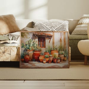 May include: A framed watercolor painting depicts a sunlit courtyard scene. Various terracotta pots filled with cacti and succulents are arranged in front of a building with a red-tiled roof and a wooden window. The artwork evokes a warm, Mediterranean aesthetic.