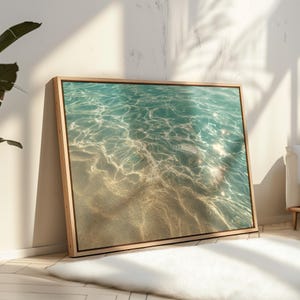 Può includere: Una stampa incorniciata con una serena scena oceanica. L'opera cattura il gioco della luce solare sulla superficie dell'acqua, creando riflessi scintillanti. Il fondale sabbioso è visibile sotto l'acqua limpida e turchese. La cornice è in legno chiaro.
