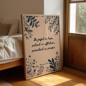 May include: Framed art print with a light pink background and navy blue floral illustrations. The print displays the text "Be joyful in hope, patient in affliction, persistent in prayer. Romans 12:12."