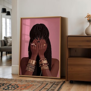 May include: A framed art print featuring a woman with dark skin and long, braided hair. Her hands, adorned with gold rings and bracelets, cover her face. The background is a solid pink, and the frame is gold.