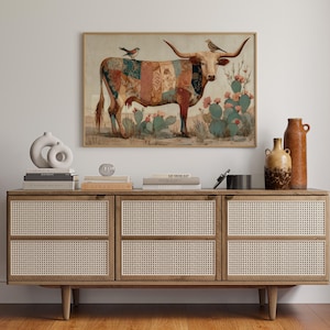 Puede incluir: Una impresión artística enmarcada que presenta una vaca longhorn con un diseño de patchwork, adornada con pájaros y ambientada en un paisaje desértico con cactus. La obra de arte se exhibe sobre un mueble de madera con paneles de puerta tejidos, complementado con jarrones y libros decorativos.