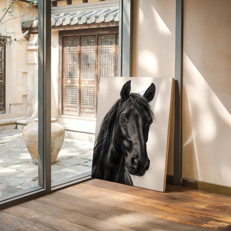 May include: A framed canvas print featuring a detailed portrait of a black horse. The horse's head and upper neck are depicted against a neutral background. The artwork is leaning against a wall in a room with a wooden floor and a view of an outdoor courtyard.