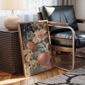 May include: A framed art print featuring a detailed illustration of coral and sea life in shades of peach, green, and cream against a dark background. The artwork is displayed on a wooden floor, leaning against a black leather chair.