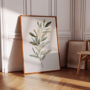 Może przedstawiać: Ramka z akwarelą przedstawiającą zieloną gałązkę oliwną z liśćmi w odcieniach zieleni i brązu. Obraz jest na białym tle i ma jasnobrązową ramę.