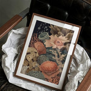 May include: A framed art print featuring a detailed illustration of a coral reef. The artwork showcases various coral formations in shades of orange, green, and beige against a dark blue background. The print is encased in a wooden frame and rests on white paper.