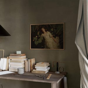 Op de afbeelding: Een ingelijst kunstwerk toont een vrouw in een witte jurk die tussen waterlelies ligt. Het kunstwerk hangt boven een houten bureau met stapels boeken, een kleine keramische beker en een schaar. Een zwarte lamp en een lichtblauw gordijn zijn ook zichtbaar.