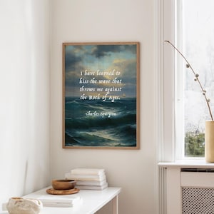 Könnte beinhalten: Gerahmter Kunstdruck mit einer Meereslandschaft und einem Zitat von Charles Spurgeon: "Ich habe gelernt, die Welle zu küssen, die mich gegen den Felsen der Ewigkeit wirft." Das Kunstwerk zeigt ein stürmisches Meer unter einem bewölkten Himmel.