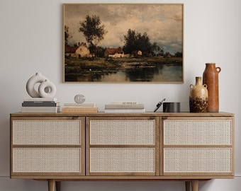 May include: A framed landscape painting depicting a serene waterside scene with houses and trees. The painting hangs above a wooden cabinet with woven cane door panels. Decorative vases and books are arranged on the cabinet's surface.