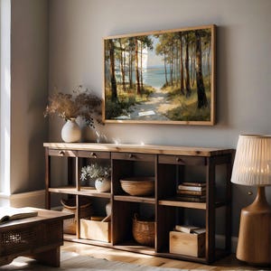 May include: A living room scene featuring a large framed landscape painting of a forest path leading to the ocean. Below the painting is a dark wood console table with open shelves, drawers, and decorative items. A wooden lamp with a cream shade sits on the right.