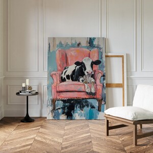 Puede incluir: Una vaca negra y blanca tumbada en un sill&oacute;n rosa. El sill&oacute;n est&aacute; delante de una pintura con pinceladas abstractas azules y blancas. La vaca mira al espectador.