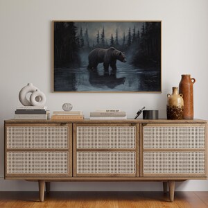 Op de afbeelding: Een ingelijst schilderij van een beer in het water, tegen een bos. Het kunstwerk hangt boven een houten kast met geweven panelen. Vazen en boeken zijn op de kast geplaatst.