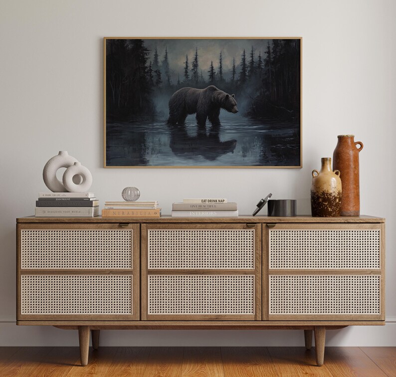 Op de afbeelding: Een ingelijst schilderij van een beer in het water, tegen een bos. Het kunstwerk hangt boven een houten kast met geweven panelen. Vazen en boeken zijn op de kast geplaatst.