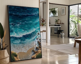 May include: A framed art print depicting an aerial view of a beach scene. The artwork features turquoise and deep blue ocean waves crashing onto a sandy shore with rocky outcroppings. The frame is black, and the artwork is displayed in a well-lit room.