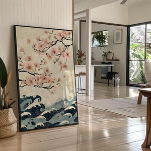 May include: A framed art print featuring a Japanese-inspired design. The artwork depicts cherry blossoms with pink flowers and dark branches, above stylized blue waves. The background is a soft beige color, and the frame is black.