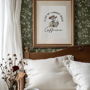 May include: A framed print with the text "A Cup of Cowboy Magic Caffeine" featuring a skeleton cowboy holding a coffee cup. The print is in a gold frame, hanging above a bed with white pillows and a wooden headboard. A vase of dried flowers is on the left.