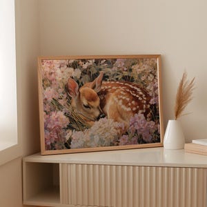May include: A framed print featuring a sleeping fawn nestled among pastel-colored flowers. The fawn has brown fur with white spots and is surrounded by white, pink, and purple blooms. The frame is a light wood color.