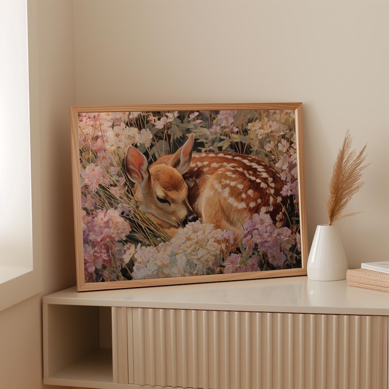 May include: A framed print featuring a sleeping fawn nestled among pastel-colored flowers. The fawn has brown fur with white spots and is surrounded by white, pink, and purple blooms. The frame is a light wood color.