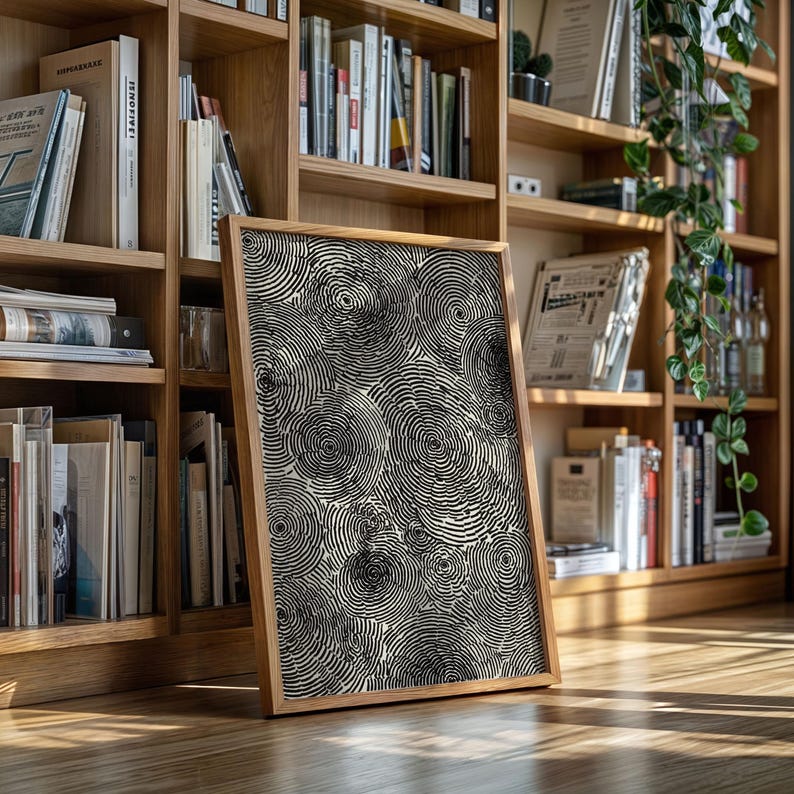 May include: A framed artwork featuring a black and white spiral pattern. The artwork is displayed on a wooden floor, leaning against a bookshelf filled with books. The frame is made of light-colored wood.