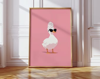 May include: A framed art print featuring a white duck wearing black sunglasses and a white towel on its head, set against a pink background. The artwork is in a gold frame, with a wooden floor and white wall in the background.
