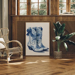 May include: A framed print featuring a pair of white and blue cowboy boots. The boots have a floral design and a blue bow. The artwork is displayed in a room with wooden paneling and a wicker chair.