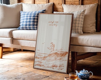 May include: A framed art print with a brown wooden frame. The print features a surfer riding a wave in a sepia tone, with the words "surf up cowboy" in cursive. The print is leaning against a beige sofa with blue and white checkered pillows.