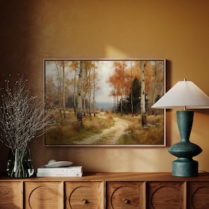 Puede incluir: Una pintura de paisaje enmarcada representa un camino que serpentea a través de un bosque de abedules con follaje otoñal. La obra de arte se exhibe sobre un mueble de madera, junto a una lámpara de mesa turquesa y un jarrón con ramas secas.
