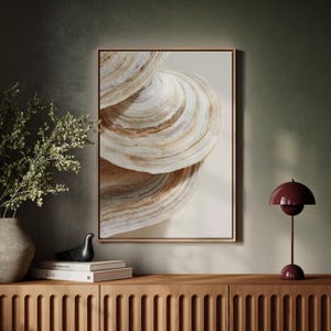 May include: A framed art print featuring a close-up of layered, textured, beige and brown abstract shapes, resembling a seashell. The artwork is displayed above a wooden cabinet, with a vase of greenery and a burgundy table lamp.