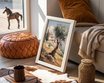 Puede incluir: Una pintura de acuarela enmarcada de un paisaje con un marco blanco. La pintura representa un árbol y un pueblo. También son visibles un puf de cuero marrón, una taza marrón y una cesta de mimbre.
